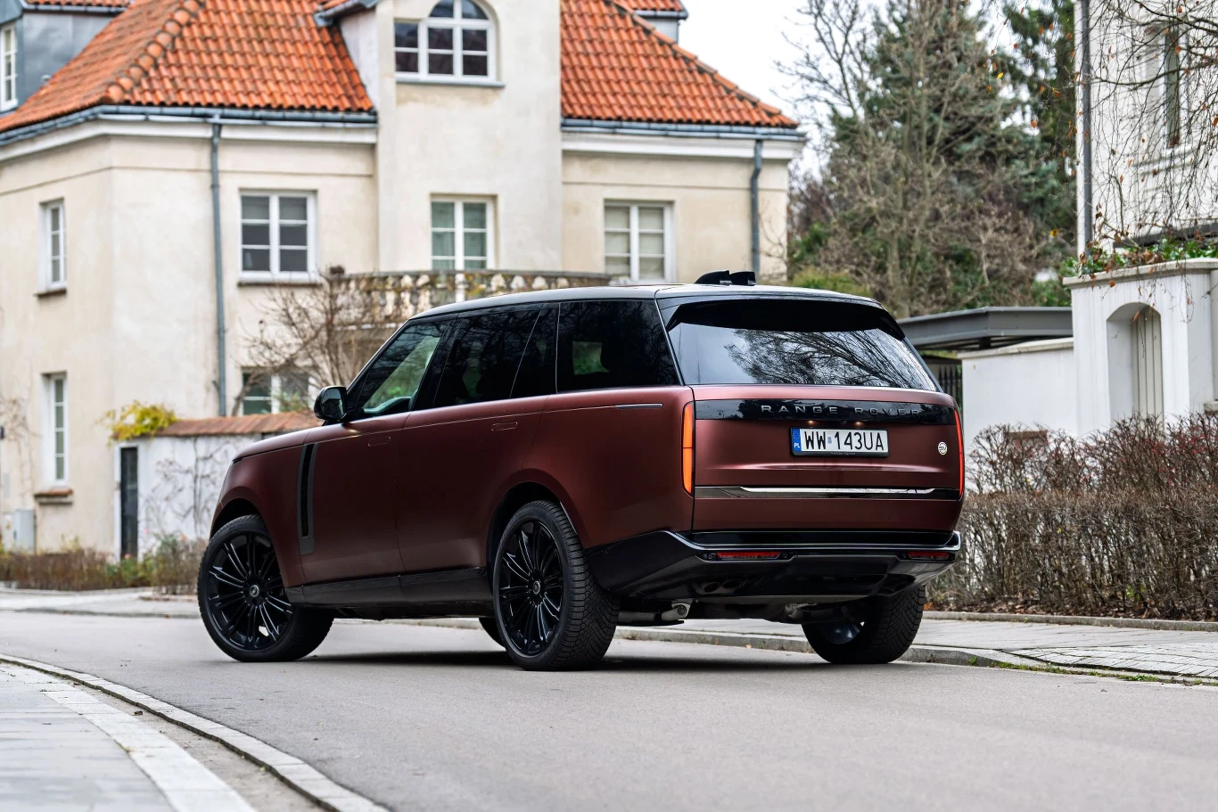 Luksusowy SUV Range Rover w matowym bordowym kolorze, zaparkowany na spokojnej, willowej ulicy, na tle eleganckiej kamienicy z czerwoną dachówką.