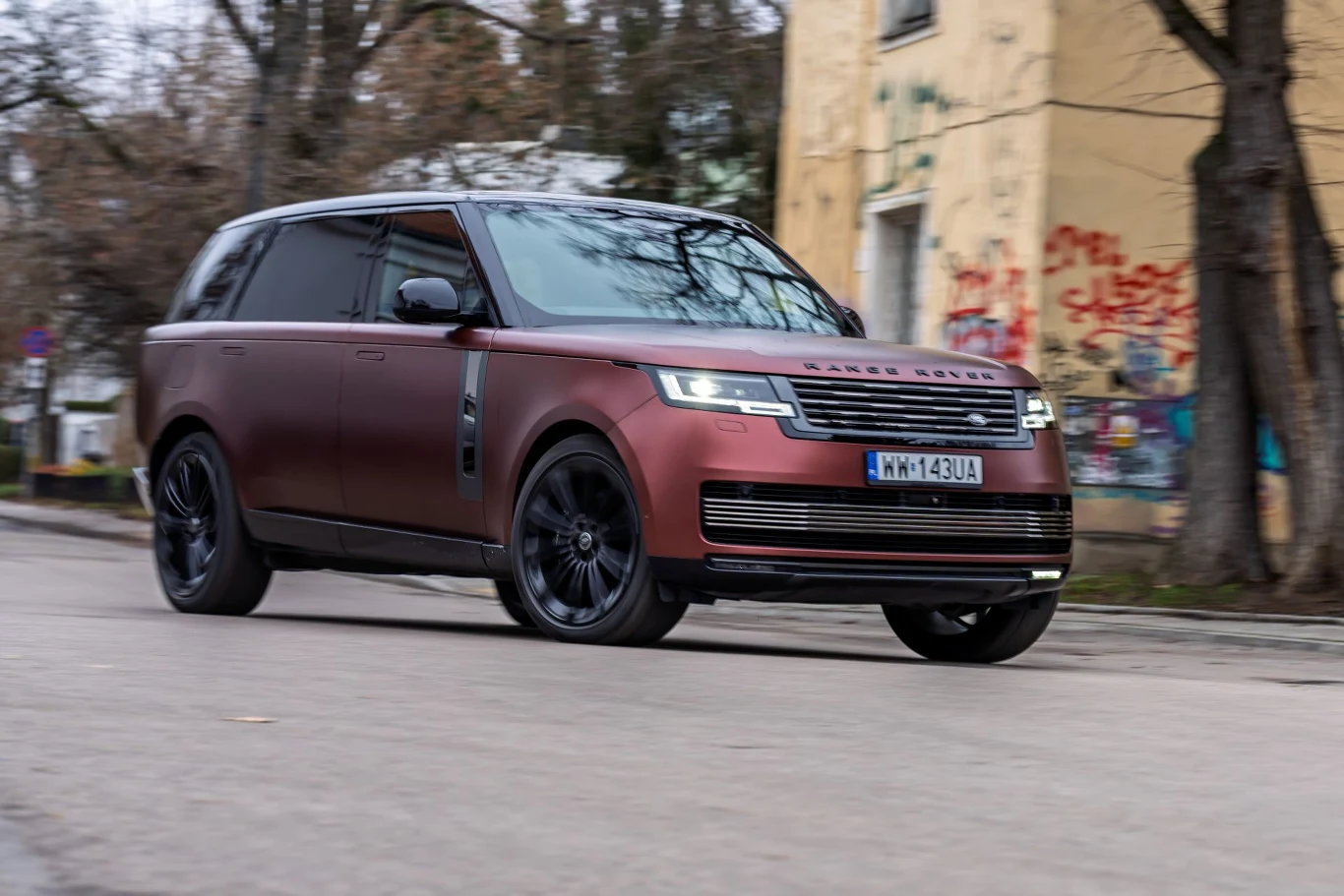 Bordowy Range Rover z czarnymi felgami porusza się miejską ulicą, na tle starego budynku pokrytego graffiti.