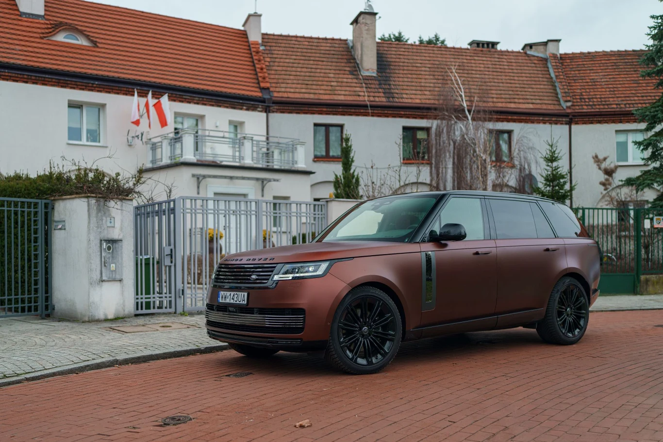 luksusowy, brązowy Range Rover zaparkowany na czerwonej kostce brukowej przed segmentem domów z białą elewacją i czerwonymi dachami, na jednym z balkonów widoczne dwie polskie flagi