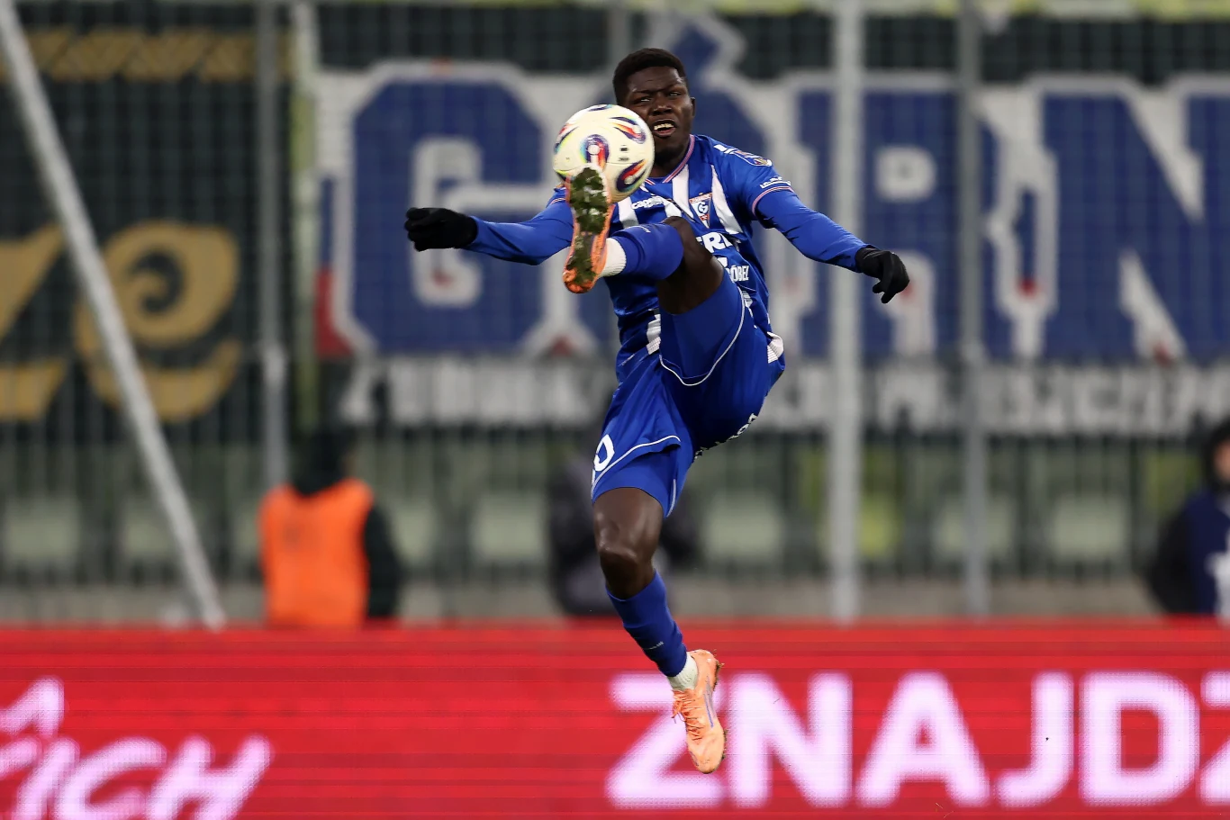 Ousmane Sow Piłkarz w niebieskim stroju wykonuje dynamiczny wykop piłki w powietrzu podczas meczu na stadionie.