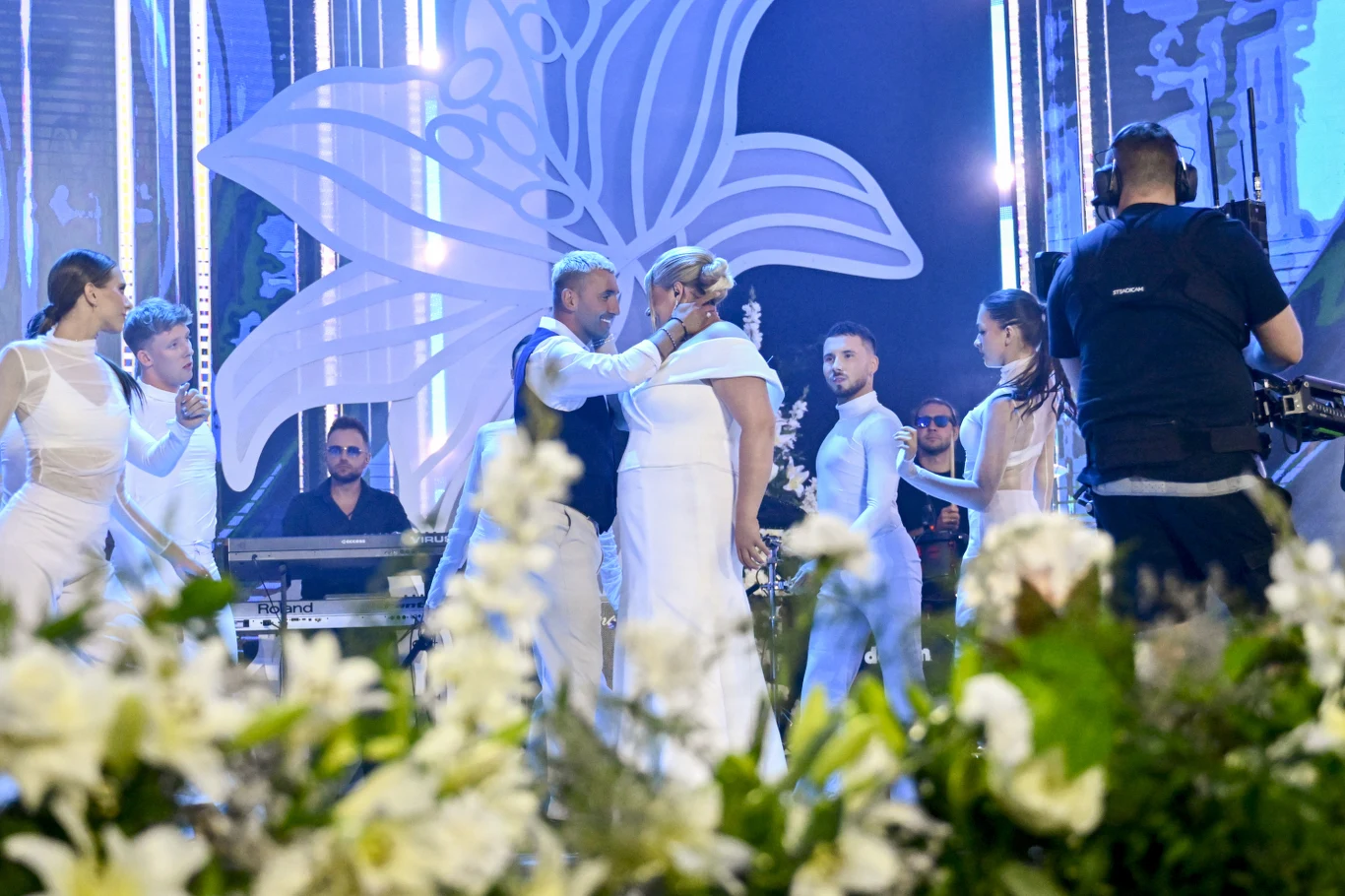 Grupa tancerzy i wokalistów ubranych na biało występuje na scenie ozdobionej dużą dekoracją w kształcie kwiatu, w otoczeniu kamerzystów i muzyków na tle jasnych świateł i kwiatów.
