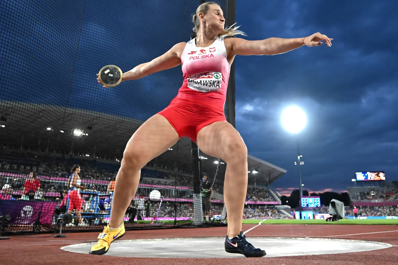 Zawodniczka w stroju reprezentacji Polski wykonuje rzut dyskiem na stadionie lekkoatletycznym podczas zawodów sportowych.