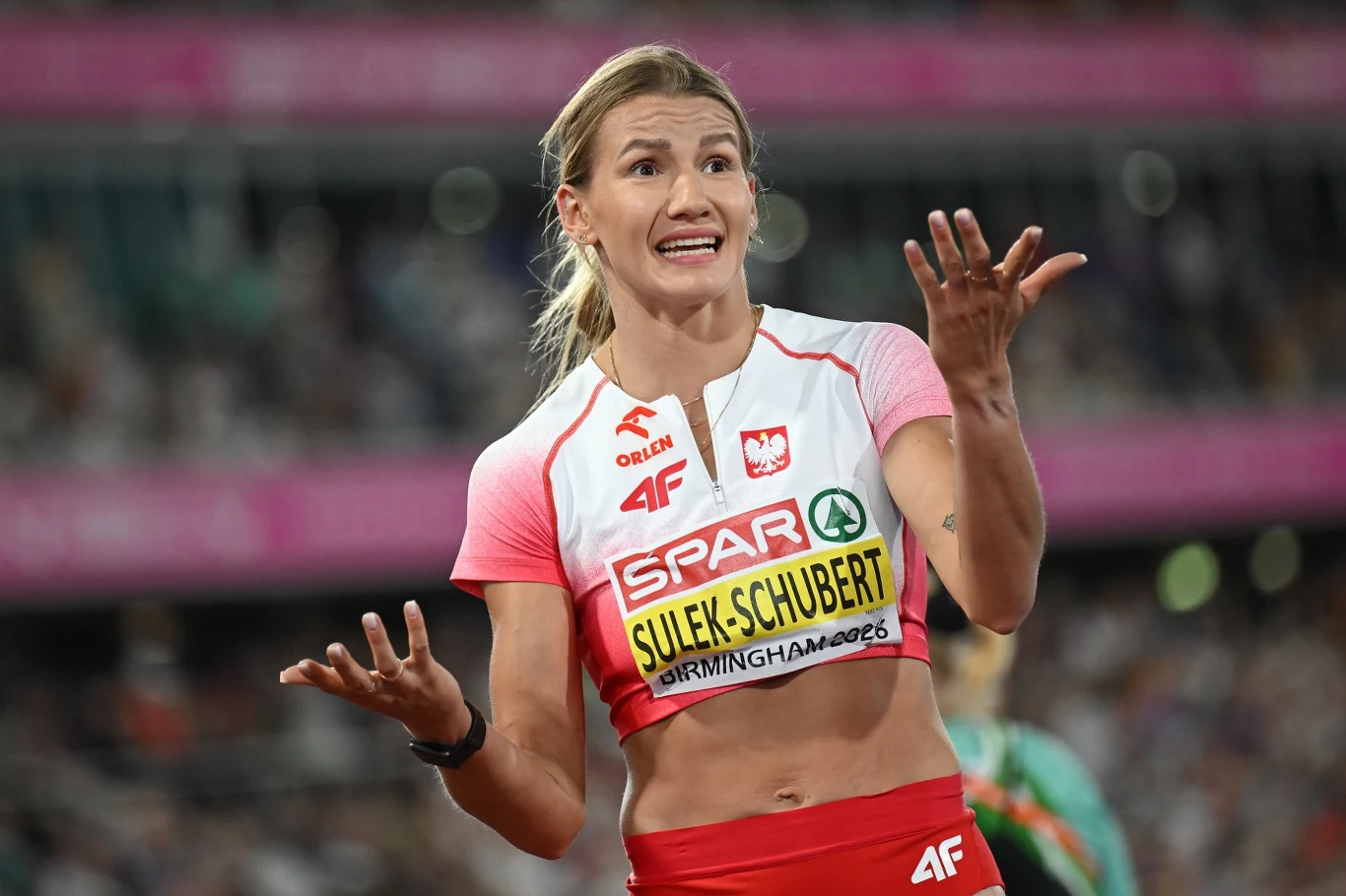 Lekkoatletka w biało-czerwonym stroju reprezentacji Polski z wyrazem zaskoczenia na twarzy, trzymająca ręce w górze z rozłożonymi dłońmi, na tle stadionu sportowego.