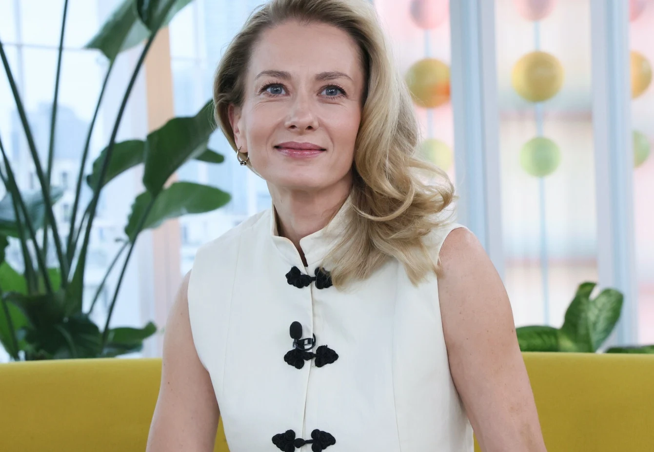 Anita Sokołowska Anita Sokołowska w białej sukni z czarnymi guzikami, siedzi na żółtej sofie w jasnym wnętrzu z roślinami.
