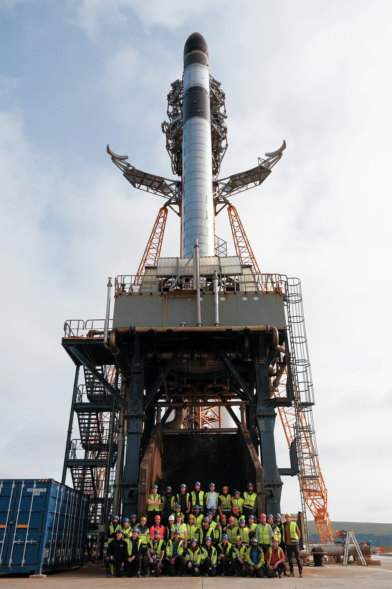 Rakieta RFA ONE Niemców z Rocket Factory Augsburg na wyrzutni w SaxaVord Spaceport. Rakieta przygotowana do startu na stanowisku, otoczona konstrukcją techniczną, przed którą stoi grupa pracowników w odblaskowych kamizelkach.