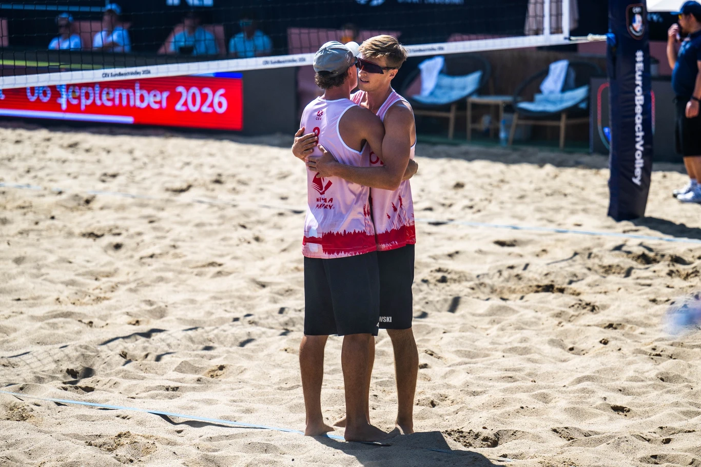 Duet Czachorowski/Krzemiński sprawił w Starych Jabłonkach pozytywną niespodziankę Dwóch siatkarzy plażowych w koszulkach sportowych obejmuje się na piasku boiska, w tle widoczne krzesła i tablica z datą wydarzenia.