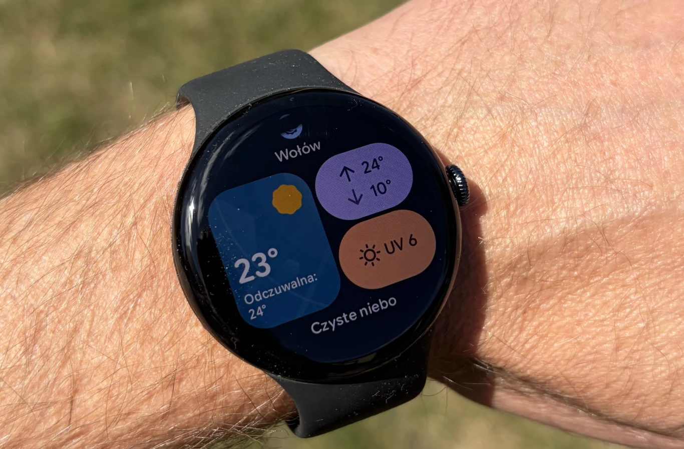 Google Pixel Watch 5 ma jasny ekran, który pozwala na wygodne korzystanie nawet w pełnym nasłonecznieniu. Czarny smartwatch na nadgarstku wyświetlający pogodę z temperaturą 23°C, odczuwalną 24°C, informacją o czystym niebie, wskaźnikiem UV na poziomie 6 oraz prognozą temperatury dla Wołowa.