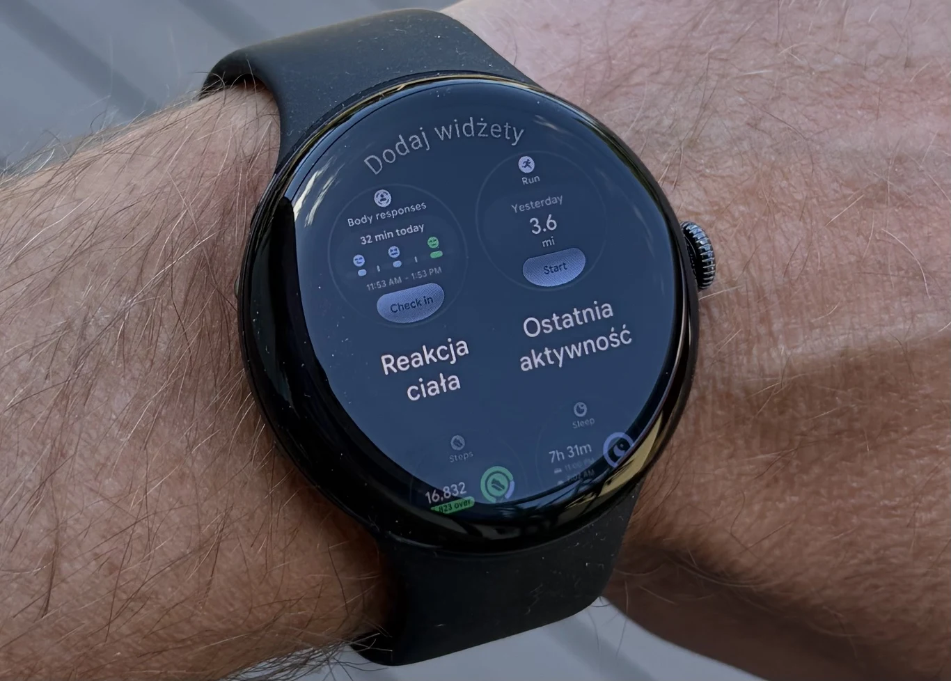 Jedną z nowości w Wear OS 7 jest wsparcie dla funkcjonalnych widżetów. Czarny smartwatch na nadgarstku, na wyświetlaczu widoczne opcje monitorowania reakcji ciała oraz ostatniej aktywności fizycznej, a także liczba wykonanych kroków i czas trwania aktywności.