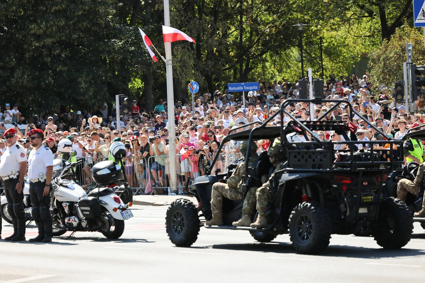Żołnierze przed defiladą wojskową na warszawskiej Wisłostradzie Żołnierze w terenowym pojeździe wojskowym oraz funkcjonariusze służb stojący obok motocykla na tle tłumu zgromadzonych widzów i powiewającej biało-czerwonej flagi.