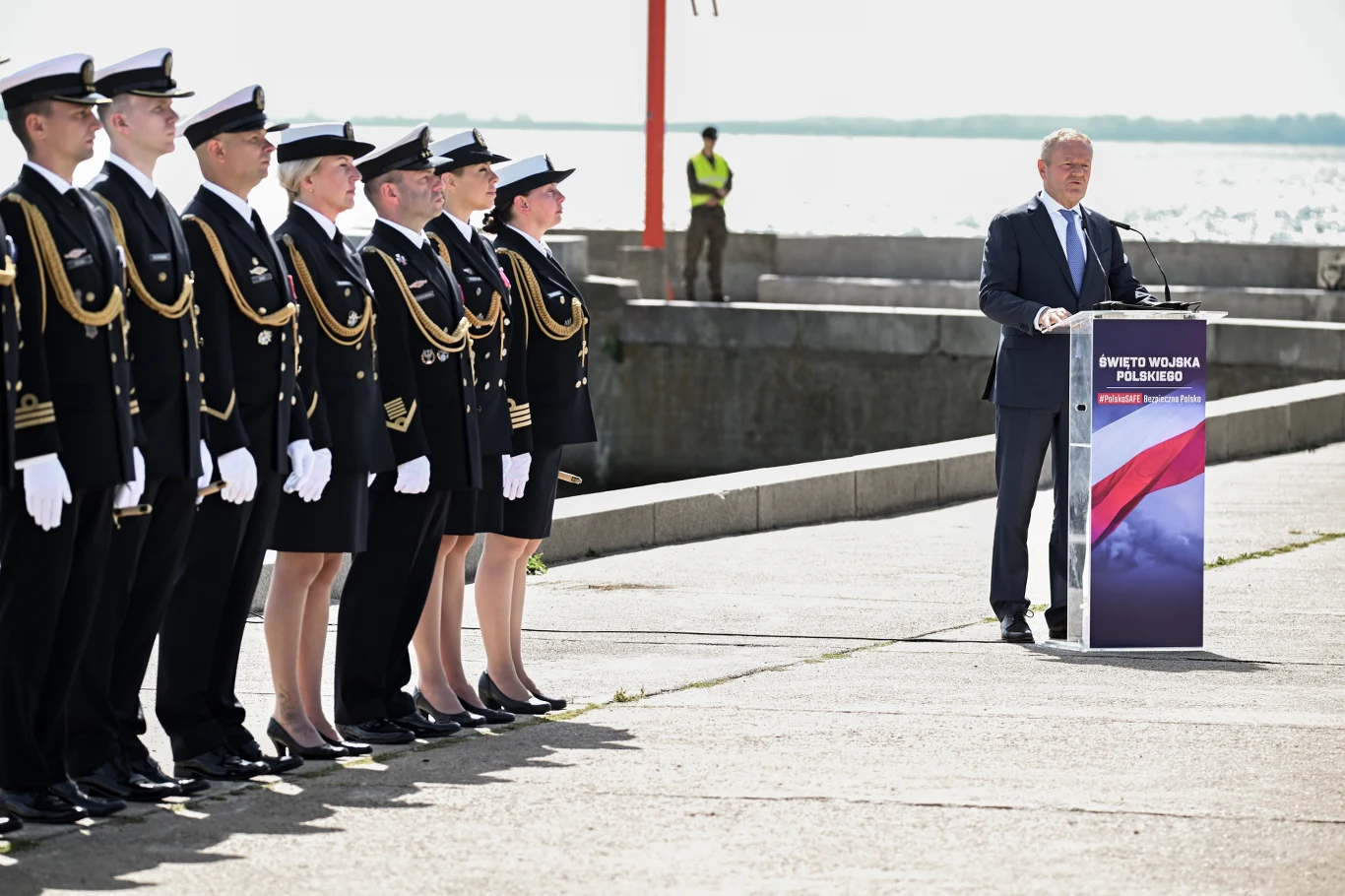 Premier Donald Tusk przemawia w Gdyni Grupa marynarzy w galowych mundurach stoi w szeregu naprzeciw mężczyzny przemawiającego przy mównicy ozdobionej biało-czerwoną flagą podczas oficjalnej uroczystości nad wodą.