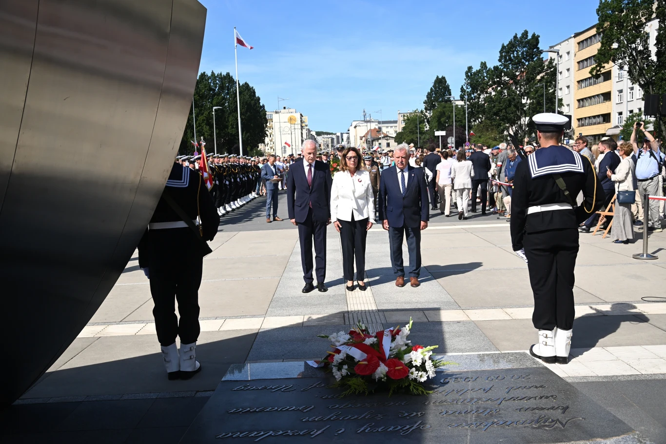 Marszałek Senatu Małgorzata Kidawa-Błońska (P) podczas złożenia kwiatów przed pomnikiem Polska Morska w Gdyni Trzy osoby stoją przed pomnikiem złożonego wieńca, w otoczeniu wartowników w mundurach i tłumu uczestników uroczystości na tle miejskiej zabudowy.