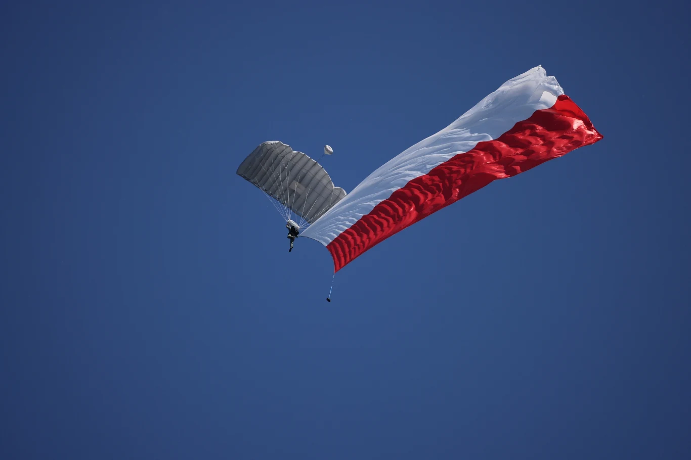 Skoczek spadochronowy z polską flagą podczas defilady wojskowej na warszawskiej Wisłostradzie Paralotniarz unoszący się na tle bezchmurnego, niebieskiego nieba z dużą, powiewającą biało-czerwoną flagą.