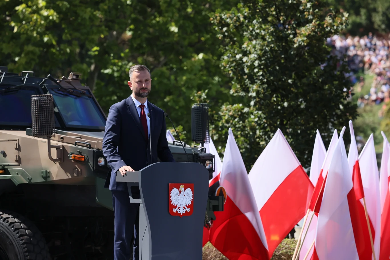 Wicepremier, minister obrony narodowej Władysław Kosiniak-Kamysz przemawia w Warszawie Mężczyzna w garniturze przemawia przy mównicy z godłem Polski, otoczony biało-czerwonymi flagami, obok stoi wojskowy pojazd opancerzony.