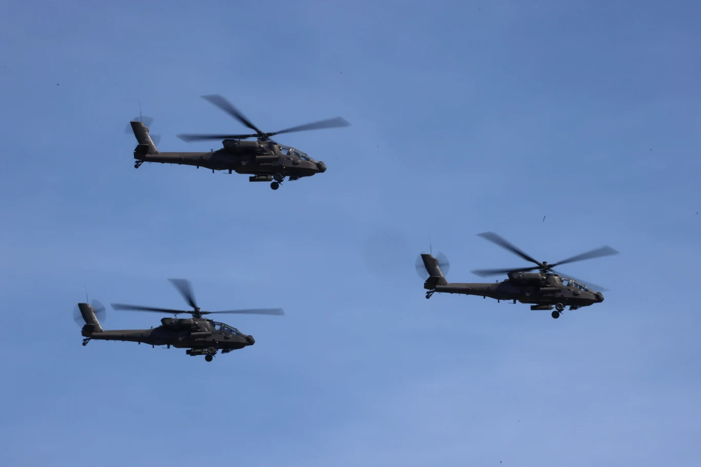 Śmigłowce AH-64 Apache podczas defilady wojskowej na warszawskiej Wisłostradzie Trzy wojskowe śmigłowce bojowe lecące w formacji na tle bezchmurnego nieba.