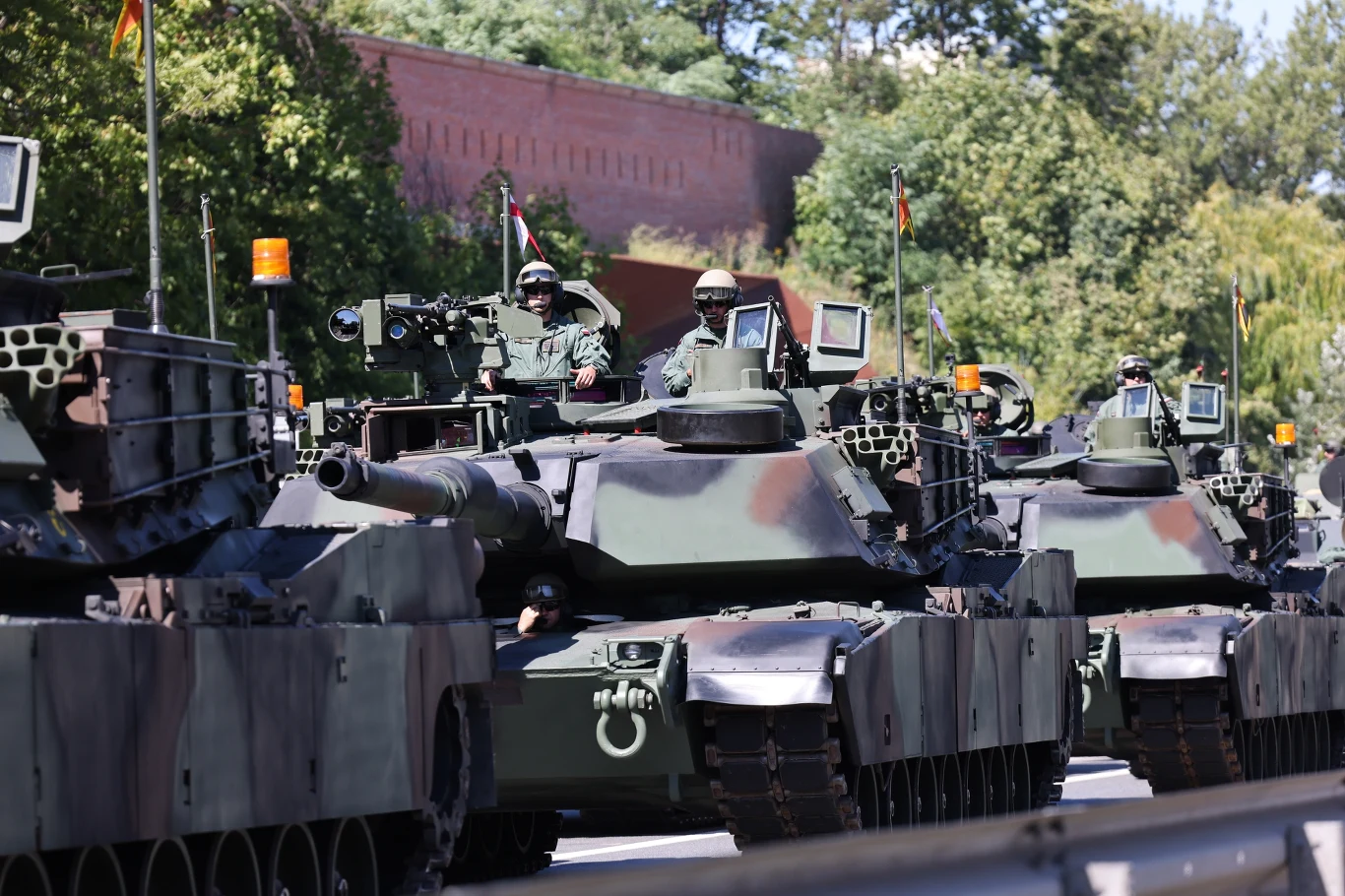 Czołgi M1 Abrams przed defiladą wojskową na warszawskiej Wisłostradzie Kolumna nowoczesnych czołgów bojowych z żołnierzami w hełmach jadącymi przez ulicę, otoczona zielenią i fragmentem ceglanych murów w tle.
