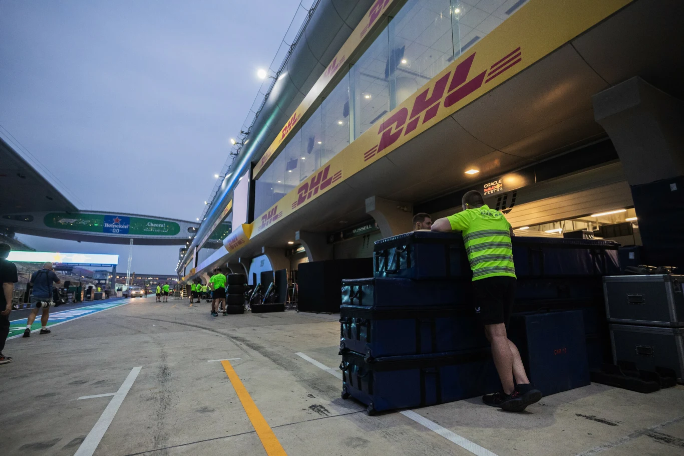 DHL ma zamiar używać morskiego transportu żeglowego Mężczyzna w odblaskowej kamizelce oparty o stos dużych plastikowych skrzyń na tle boksów serwisowych z logo DHL, w otoczeniu kilku innych pracowników przy torze wyścigowym.