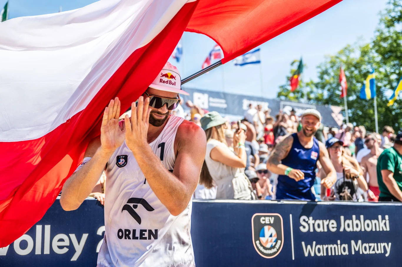Siatkarz plażowy z numerem 11 na koszulce i czapką z logo Red Bulla unosi ręce, trzymając się dużej biało-czerwonej flagi, otoczony przez kibiców na tle baneru turnieju w Starych Jabłonkach.