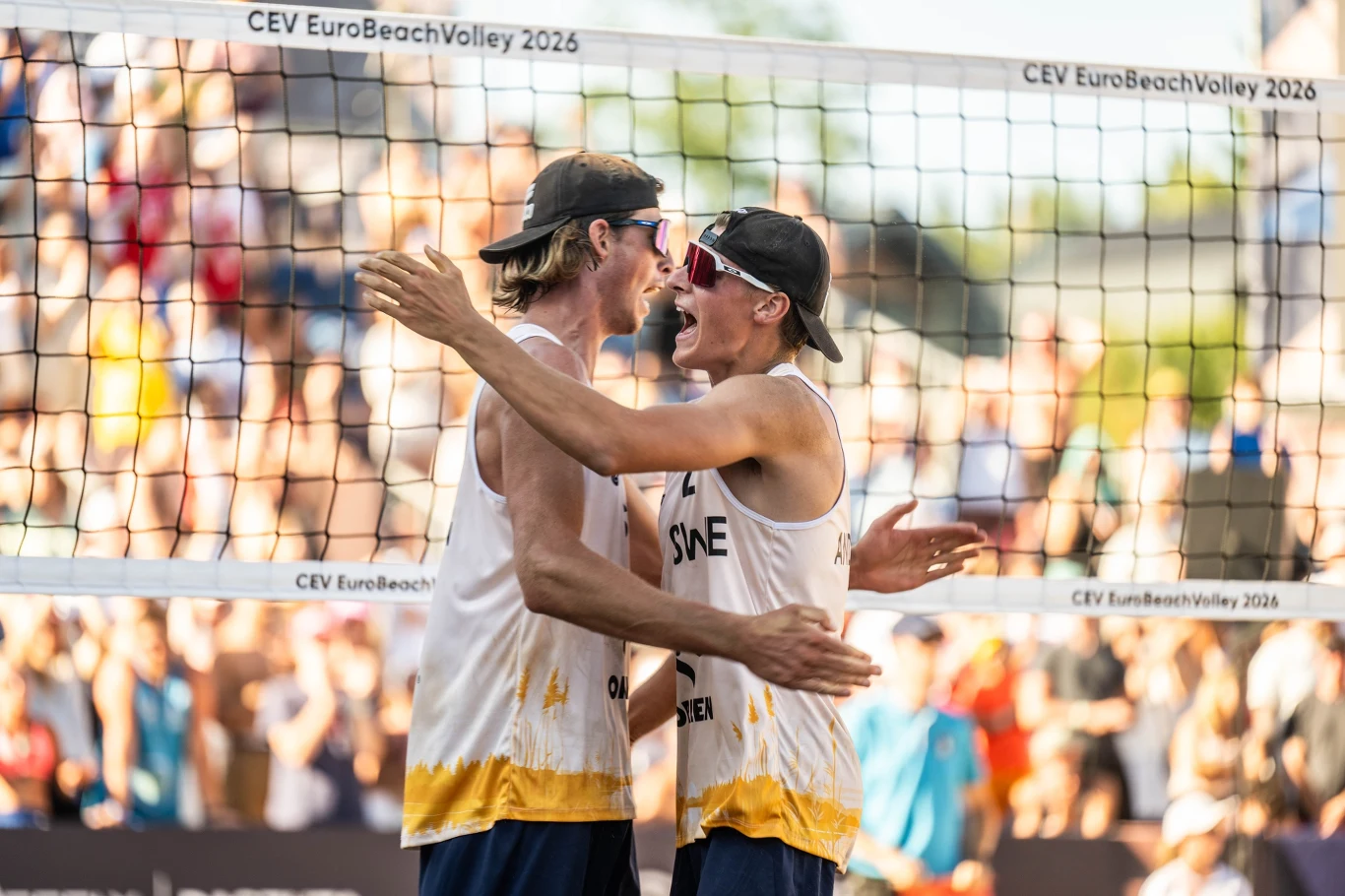 Dwóch zawodników w stroju sportowym z napisem SUE ściska się na boisku do siatkówki plażowej podczas turnieju CEV EuroBeachVolley 2026, w tle rozmyta publiczność na trybunach.