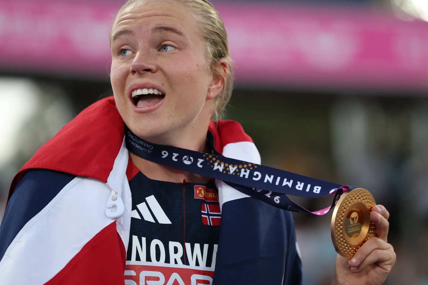 Sportsmenka owinięta flagą Norwegii z uśmiechem prezentuje złoty medal, trzymając go w dłoni podczas ceremonii dekoracji.