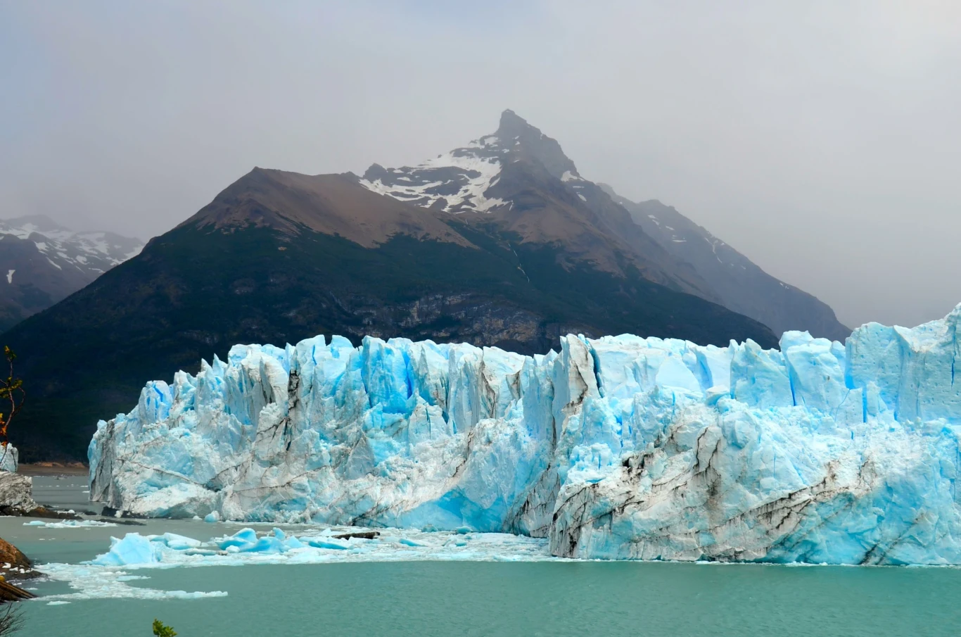 Niezwykły lodowiec Perito Moreno. Inne topnieją, a ten rośnie. Lodowiec Perito Moreno z jasnoniebieskimi szczelinami nad turkusową wodą, w tle skaliste góry z ośnieżonym szczytem.