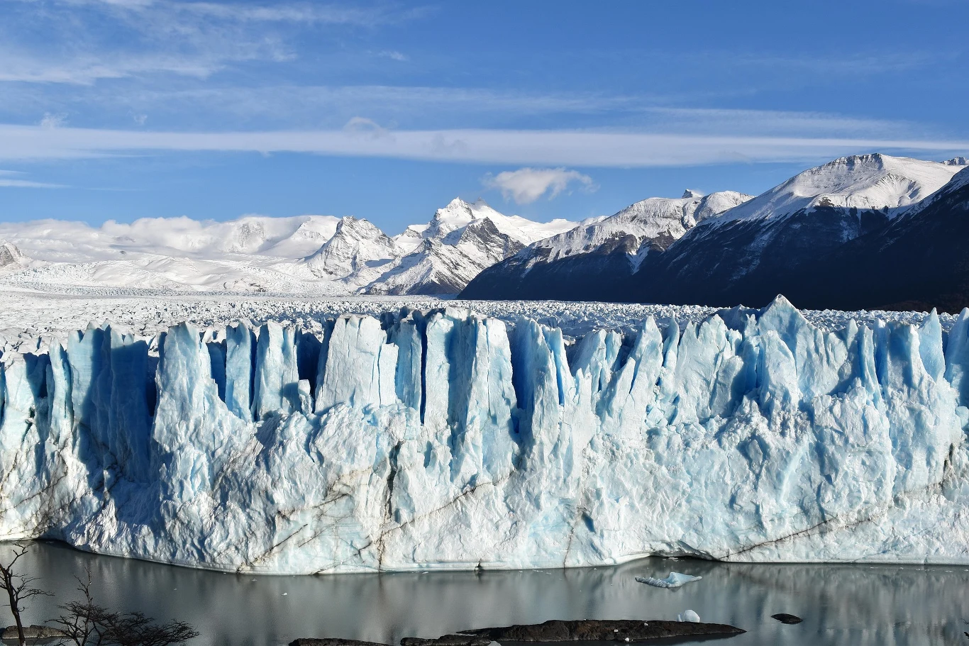 Lodowiec Perito Moreno wznosi się na około 70 metrów nad powierzchnią jeziora. Imponująca ściana lodowca z ostrymi, błękitnymi formacjami lodowymi wznosząca się nad ciemną wodą jeziora, w tle ośnieżone górskie szczyty pod błękitnym niebem.