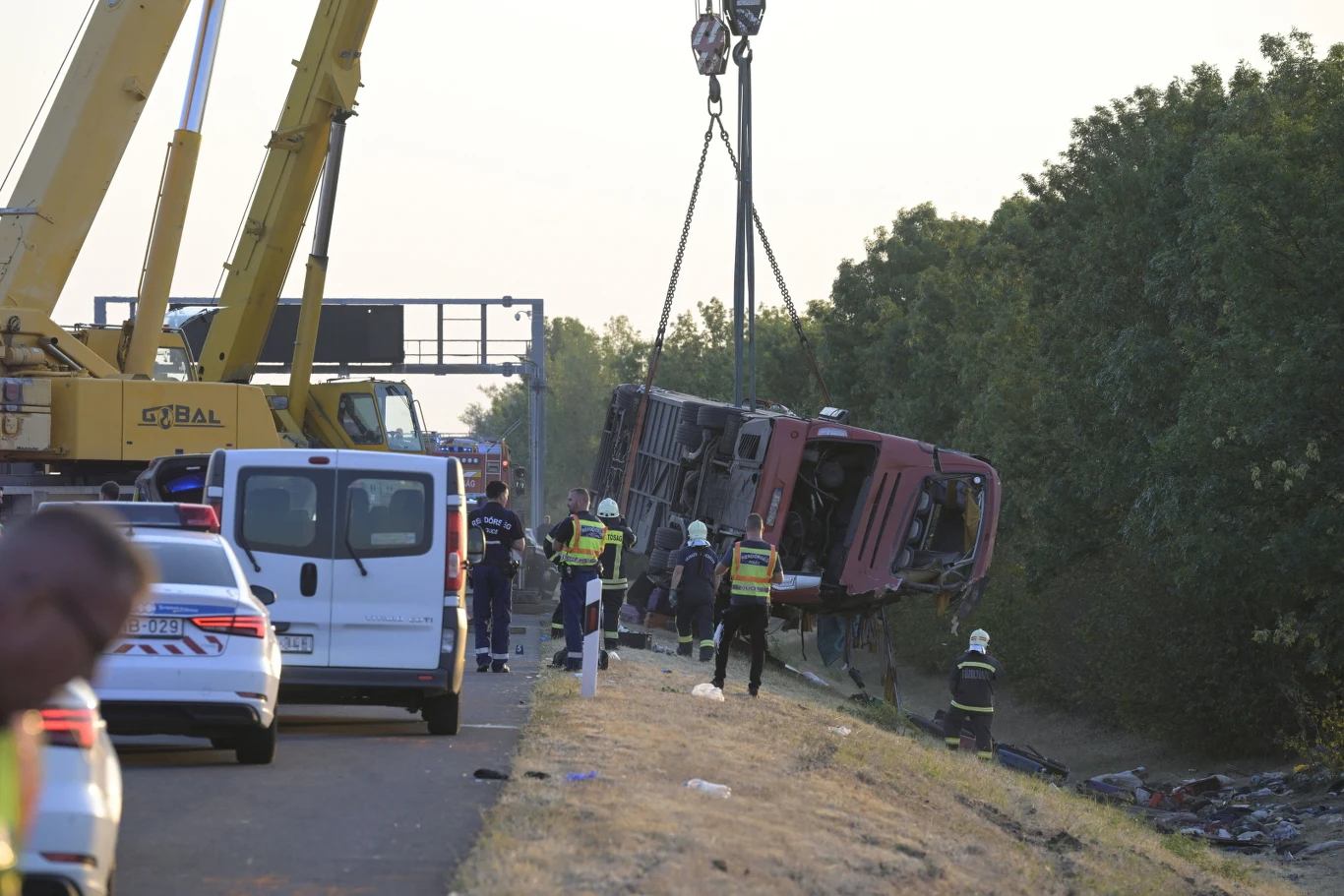 Służby ratunkowe i ciężki sprzęt unoszą przewrócony autobus przy pomocy dźwigu na poboczu drogi, w pobliżu kilka radiowozów i funkcjonariusze zabezpieczający miejsce zdarzenia.