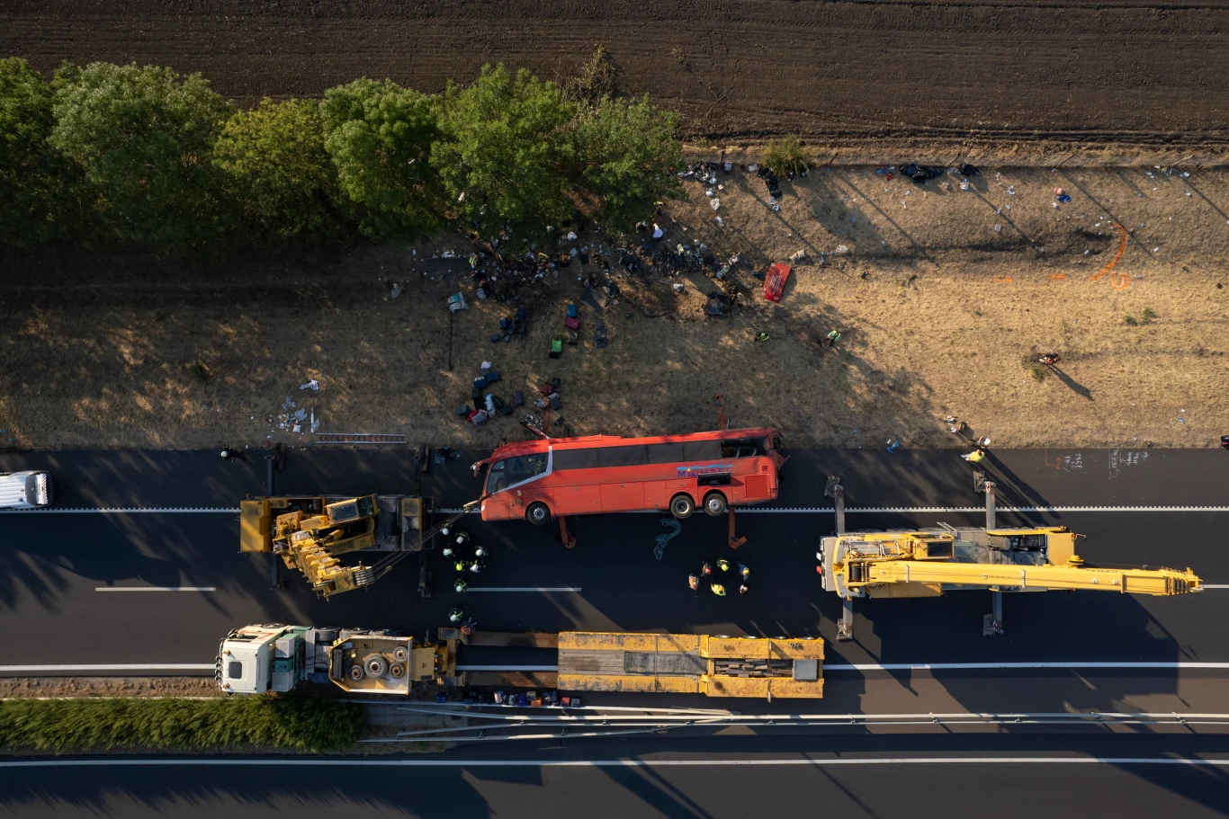 Autobus leżący na boku poza jezdnią oraz dwa żółte dźwigi i ciężarówka ustawione na drodze, wokół których pracują ratownicy i służby przy akcji ratunkowej.