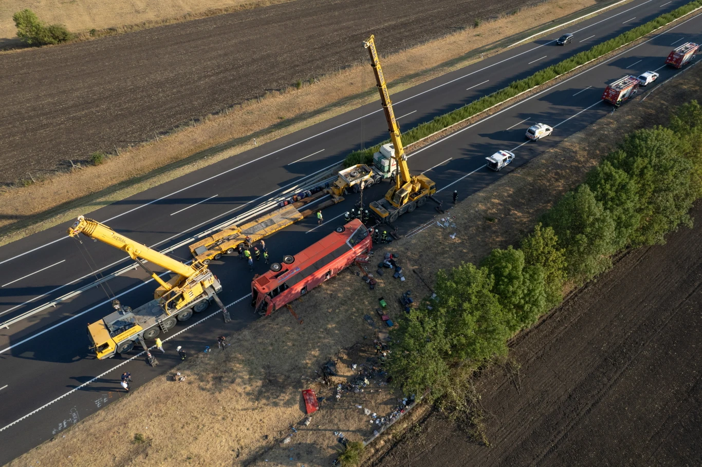 Dwa dźwigi podnoszą przewrócony czerwony autokar leżący na skraju drogi szybkiego ruchu, wokół pracują służby ratunkowe, a ruch na jezdni jest wstrzymany przez liczne wozy strażackie i policyjne.