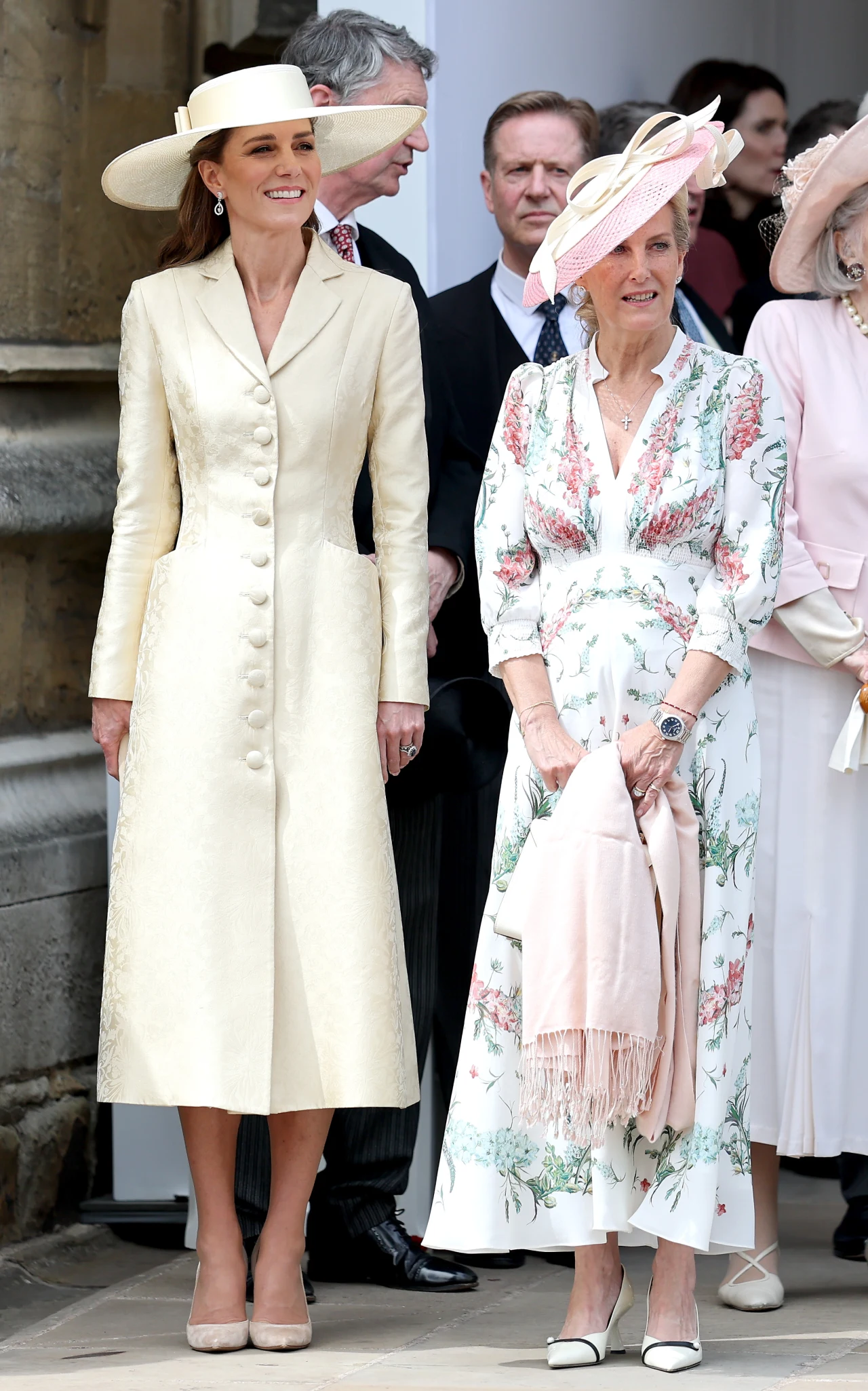Podczas gdy Kate wyznacza trendy wśród 40-latek, Zofia może inspirować kobiety po 60-tce Dwie elegancko ubrane kobiety w kremowych, jasnych kapeluszach, z czego jedna ma długi, kremowy płaszcz, a druga sukienkę w kwiaty oraz narzucony na ręce jasny szal, stoją obok siebie i uśmiechają się.