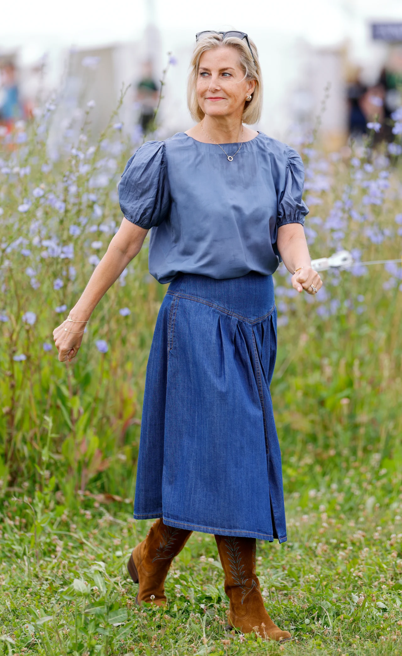 Denim i brązowe zamszowe botki to kwintesencja codziennego stylu księżnej, który jest tak bezpretensjonalny, że może być naśladowany przez każdą kobietę Kobieta o blond włosach ubrana w niebieską bluzkę z bufiastymi rękawami, długą jeansową spódnicę i zamszowe brązowe kozaki, spaceruje po łące pełnej wysokiej trawy i fioletowych kwiatów.