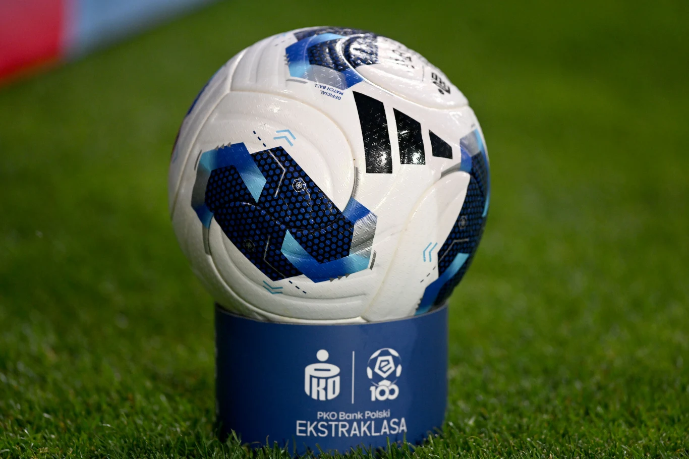 Piłka i logo PKO BP Ekstraklasy Piłka nożna z niebiesko-czarnymi wzorami ustawiona na niebieskim stojaku z logo PKO BP Ekstraklasa, umieszczona na murawie boiska.