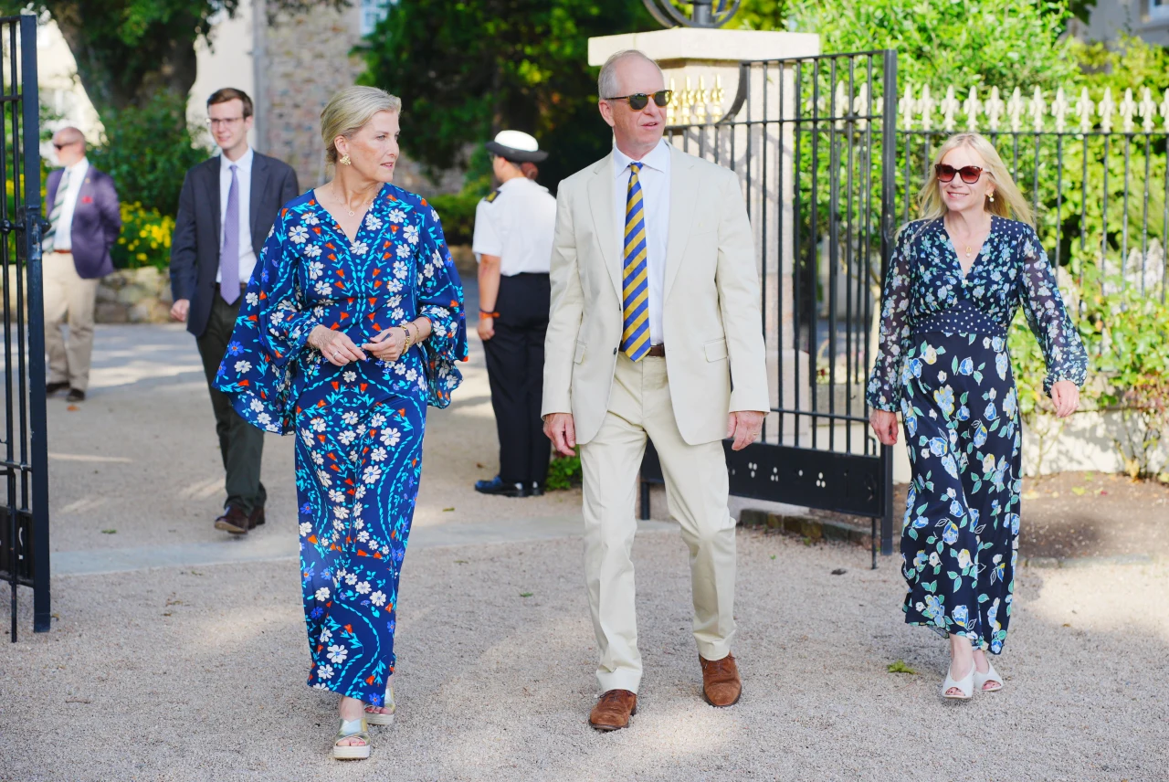Jej sukienki są często wyraziste, z efektowymi wzorami Trzy elegancko ubrane osoby idą razem, dwie kobiety w kwiecistych sukienkach i mężczyzna w jasnym garniturze z krawatem w paski, otoczone ogrodzeniem i zielenią.