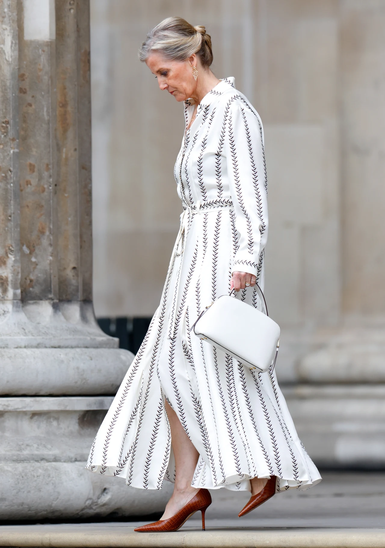 Czy zainspirujecie się stylem księżnej Zofii? Elegancka kobieta w białej sukience w geometryczne wzory, z białą torebką i brązowymi szpilkami, idzie przy kamiennych kolumnach.
