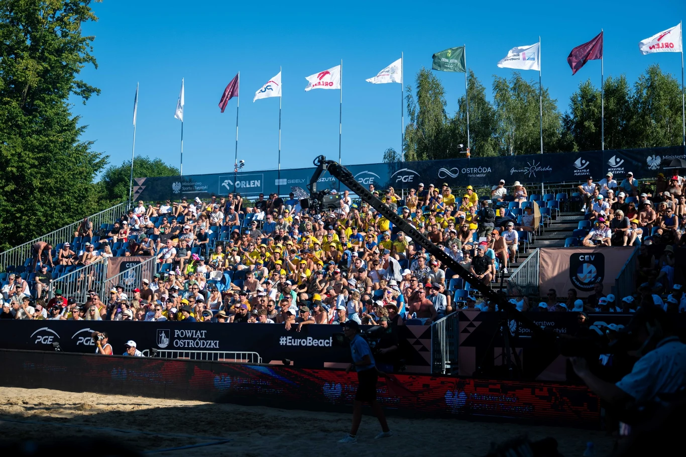 Turniej w Starych Jabłonkach trwa od środy Tłum kibiców zgromadzony na trybunach podczas turnieju siatkówki plażowej, na pierwszym planie ramię kamery telewizyjnej skierowane w stronę boiska, w tle różnokolorowe flagi powiewające nad głowami widzów.