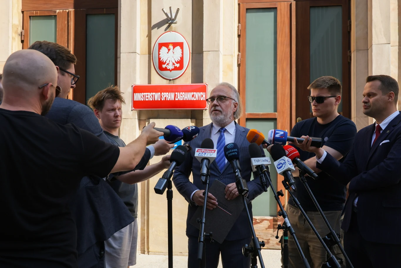 Andrzej Osiak, Naczelnik Wydziału Pomocy Konsularnej i Kryzysów Konsularnych MSZ Mężczyzna w garniturze udziela wywiadu przed budynkiem Ministerstwa Spraw Zagranicznych, otoczony przez dziennikarzy trzymających mikrofony i kamery, w tle widoczne godło Polski i czerwona tablica urzędowa.