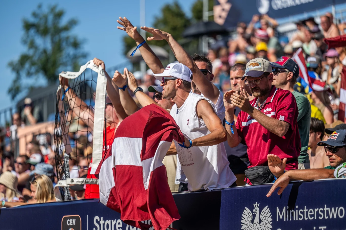 Łotewskie siatkarki przyciągnęły na trybuny wielu kibiców ze swojego kraju Grupa entuzjastycznych kibiców dopingujących podczas wydarzenia sportowego, z przodu mężczyzna trzymający flagę Łotwy, wszyscy zwróceni w stronę boiska.