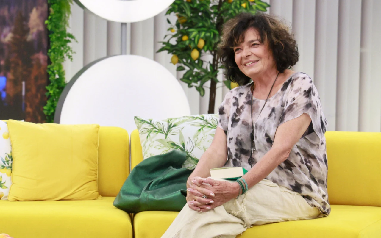 Katarzyna Grochola w "halo tu polsat" Uśmiechnięta kobieta z krótkimi, kręconymi włosami siedząca na żółtej sofie, w tle dekoracyjne poduszki i roślina cytrusowa.