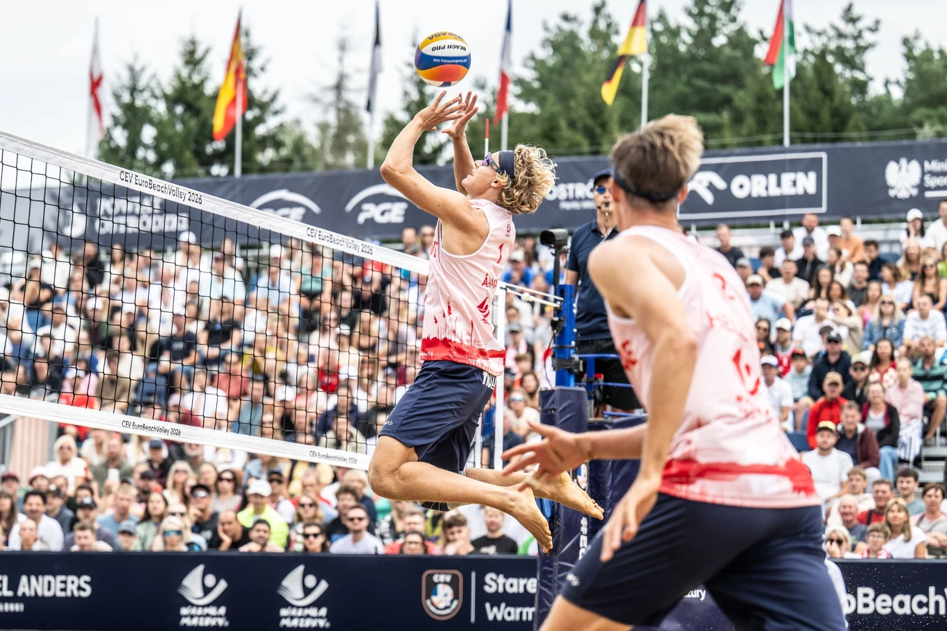 Dwóch siatkarzy plażowych w biało-czerwonych strojach wykonuje dynamiczną akcję przy siatce, jeden z nich wyskakuje do ataku piłki, a za nimi widoczna jest liczna publiczność oraz bandy reklamowe.