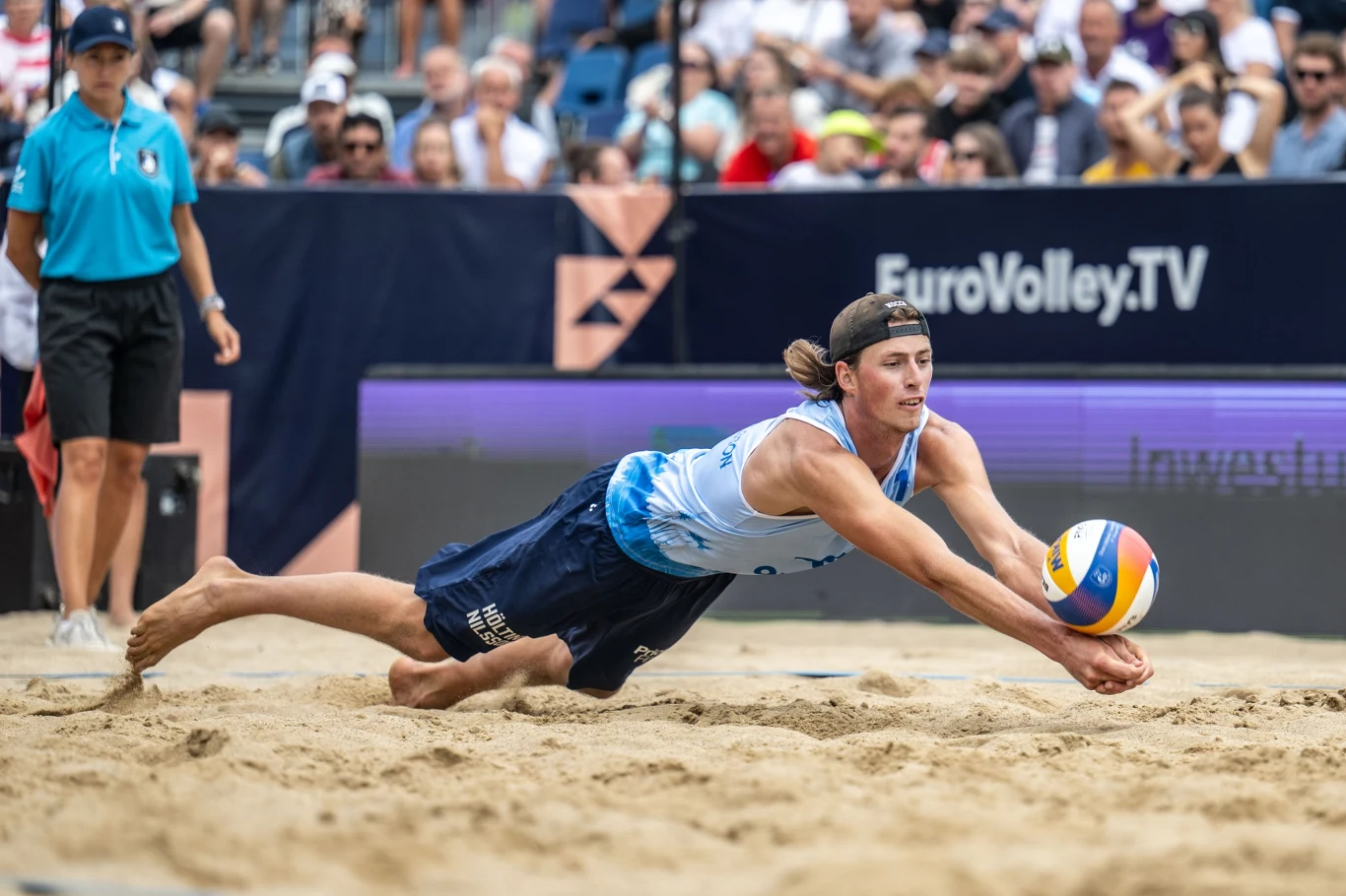 Jacob Holting Nilsson Zawodnik w stroju do siatkówki plażowej wykonuje dynamiczną interwencję, próbując odbić piłkę tuż nad piaskiem podczas meczu, w tle widoczni kibice i sędzia.