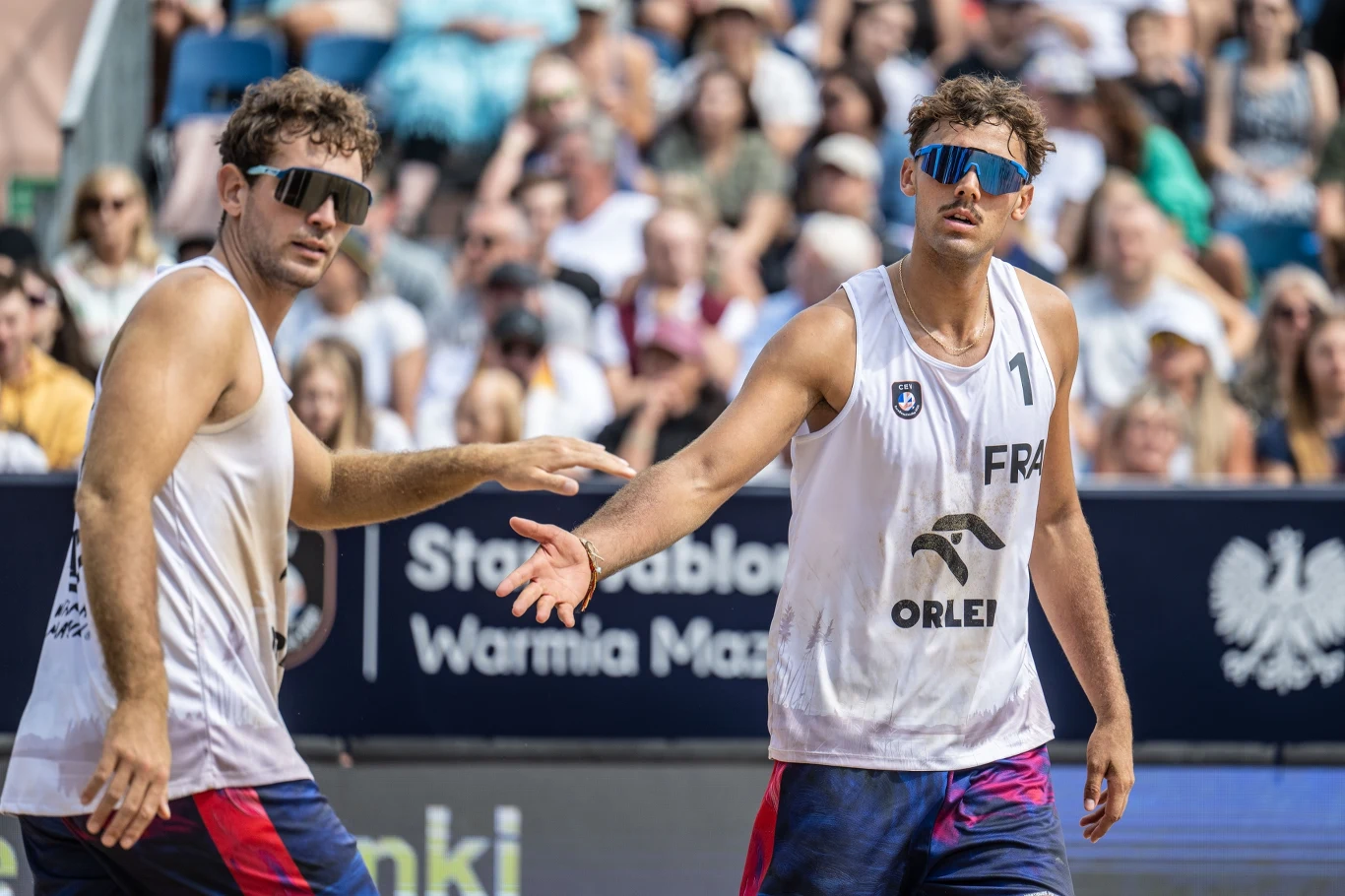 Francuski duet zdobył brązowy medal mistrzostw Europy w Starych Jabłonkach Dwóch siatkarzy plażowych w białych koszulkach i okularach przeciwsłonecznych wykonuje gest przed siebie podczas sportowych zawodów na oczach licznej publiczności.