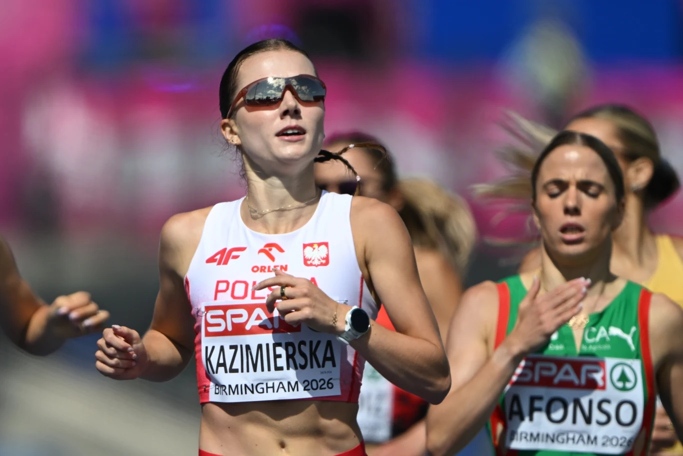 Klaudia Kazimierska Polska lekkoatletka w biało-czerwonym stroju z okularami przeciwsłonecznymi prowadzi grupę zawodniczek podczas biegu na stadionie lekkoatletycznym.