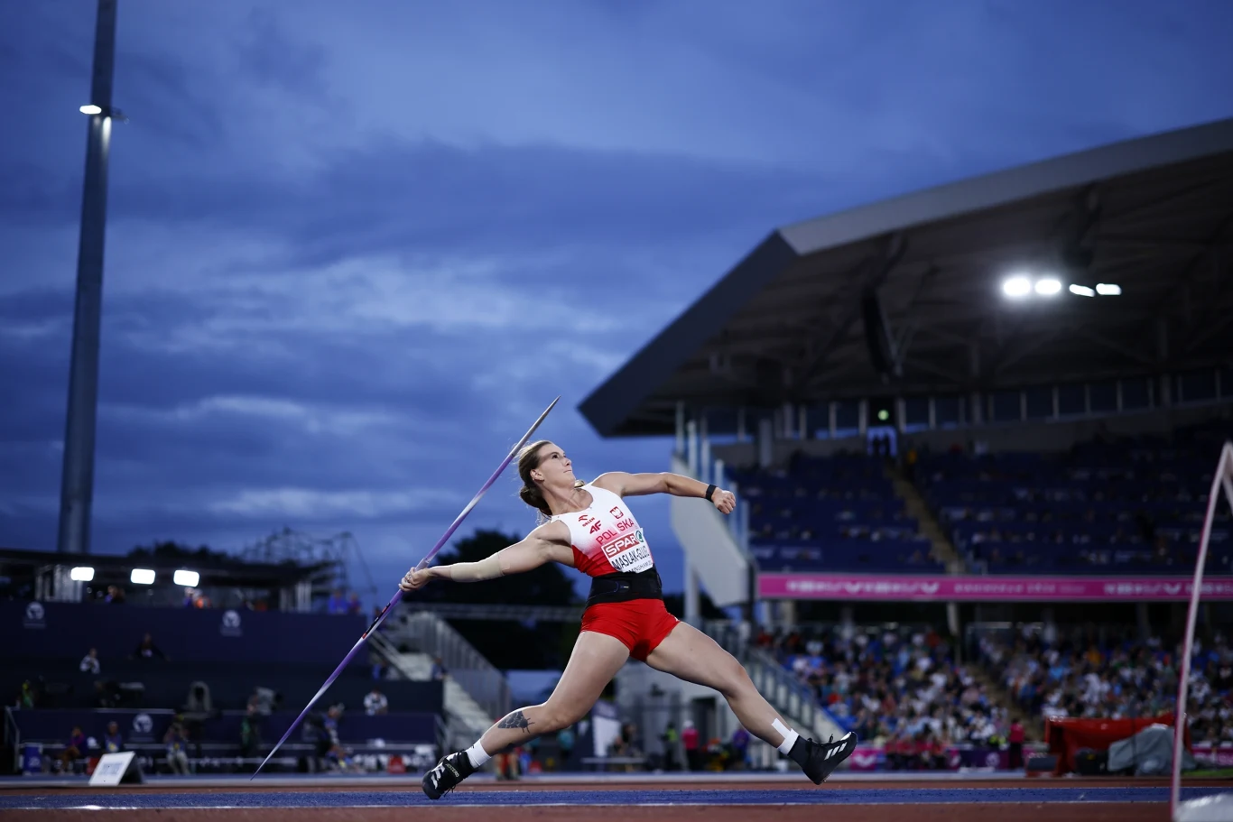Małgorzata Maślak-Glugla Zawodniczka w trakcie rzutu oszczepem na stadionie lekkoatletycznym, ubrana w czerwono-biały strój, z dynamicznym ruchem ciała podkreślającym moment wyrzutu, na tle wieczornego nieba oraz trybun wypełnionych widzami.