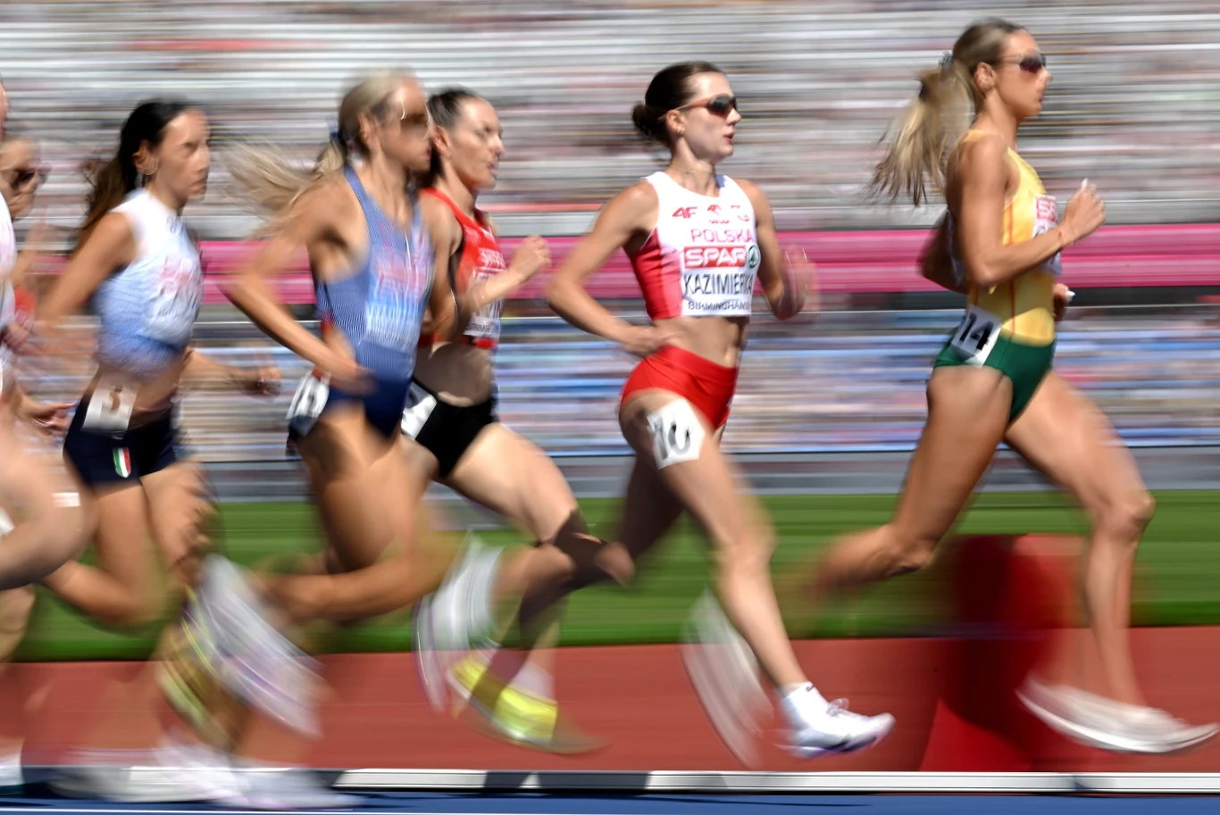 Klaudia Kazimierska Grupa zawodniczek biegnących na stadionie lekkoatletycznym podczas wyścigu na średnim dystansie, z wyraźnie zaznaczoną dynamiką ruchu i determinacją na twarzach sportowczyń.