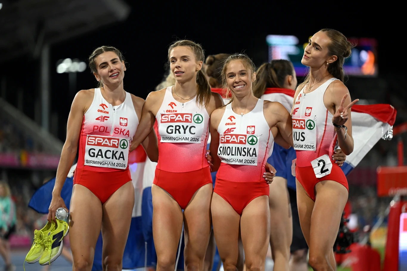 Cztery lekkoatletki w biało-czerwonych strojach reprezentacji Polski stoją tuż obok siebie na bieżni stadionu, trzymając się za ramiona, z medalami na szyjach i szerokimi uśmiechami na twarzach.