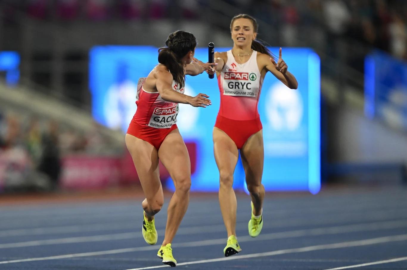 Dwie polskie lekkoatletki w czerwono-białych strojach wykonują zmianę pałeczki podczas sztafety na bieżni stadionu lekkoatletycznego.