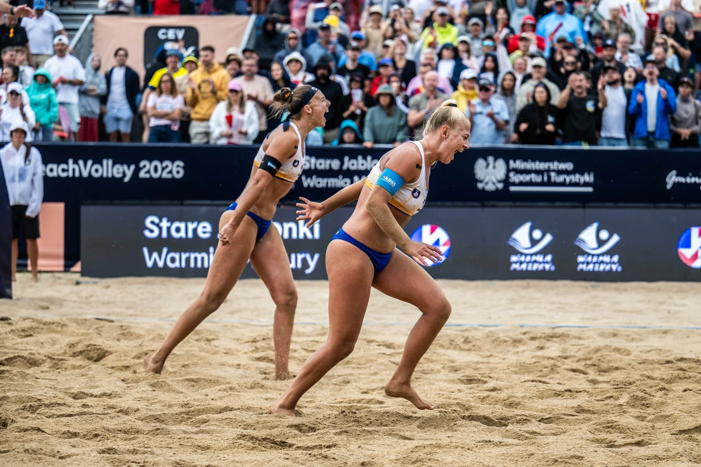Dwie zawodniczki w sportowych strojach rywalizują na piaszczystym boisku do siatkówki plażowej, w tle widoczny tłum kibiców na trybunach.