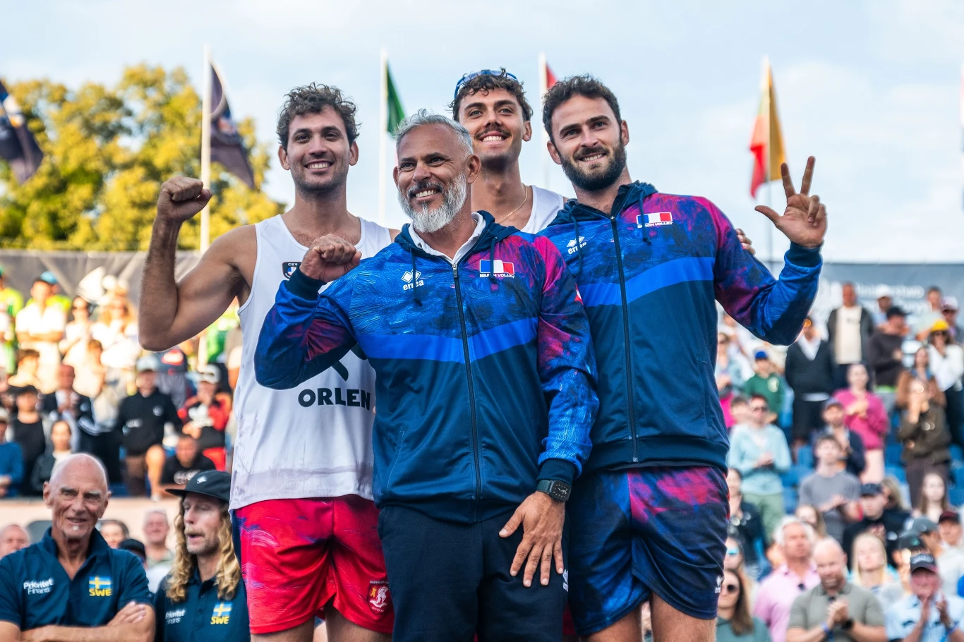 Trzech sportowców i trener w sportowych strojach pozuje na tle tłumu kibiców, prezentując medale i gesty zwycięstwa na zawodach lekkoatletycznych.