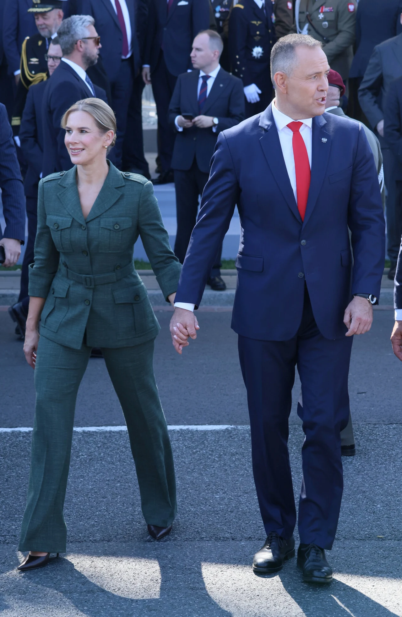 Kobieta w zielonym garniturze i mężczyzna w granatowym garniturze z czerwoną krawatą idą razem, trzymając się za ręce, wśród tłumu elegancko ubranych osób.