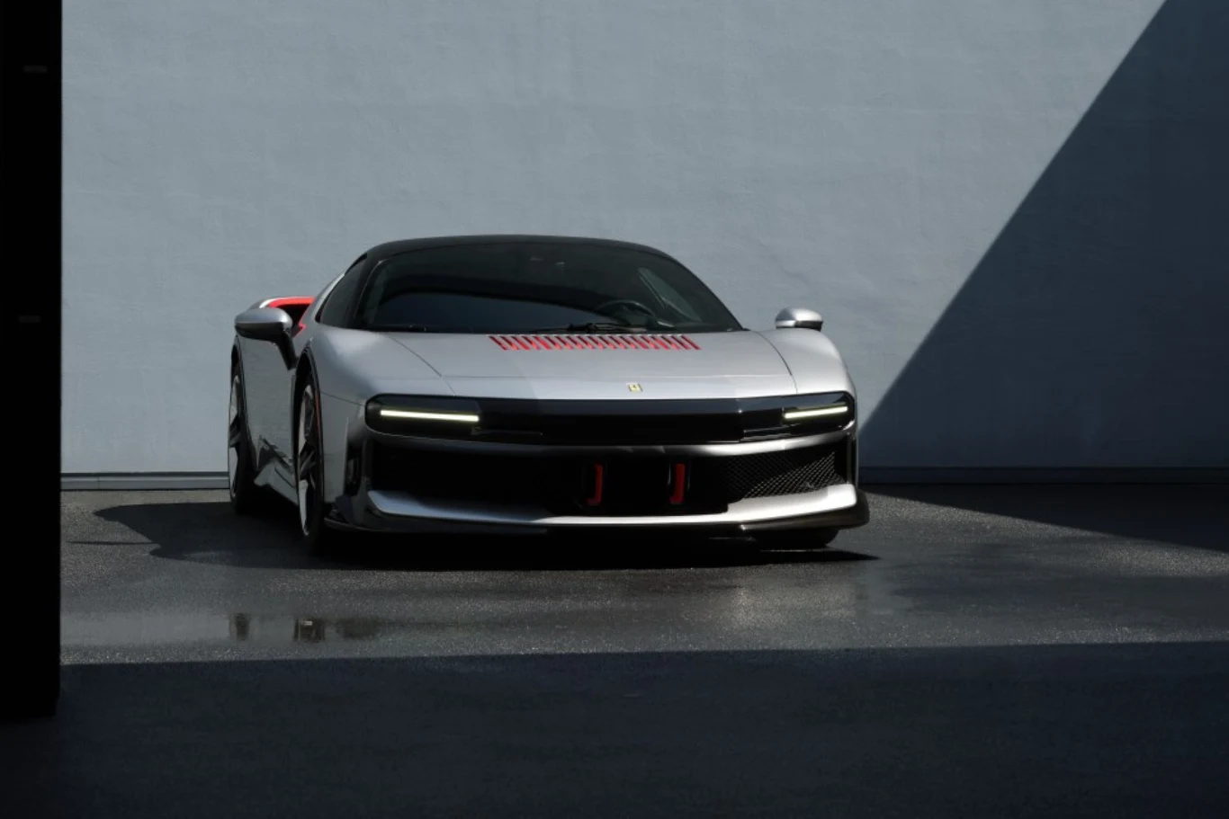 Przednie reflektory są niemal niewidoczne. Srebrne sportowe auto Ferrari z charakterystycznymi, wąskimi światłami LED oraz czerwonymi akcentami na masce i bocznych lusterkach, zaparkowane na ciemnym asfalcie przy szarej ścianie.
