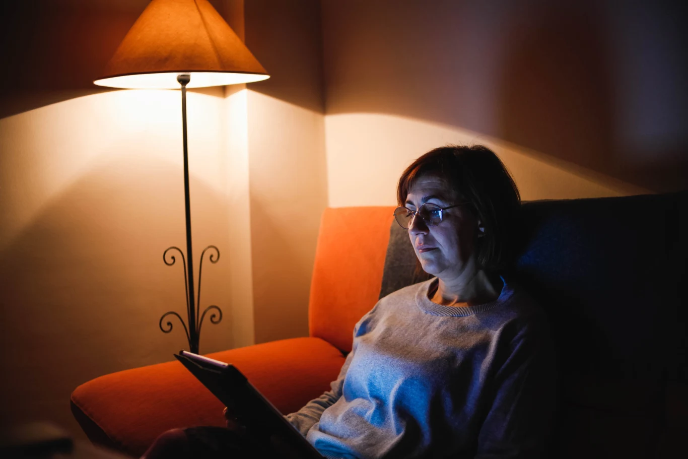 Kobieta w okularach siedzi na kanapie, trzymając tablet, oświetlona ciepłym światłem lampy stojącej obok.